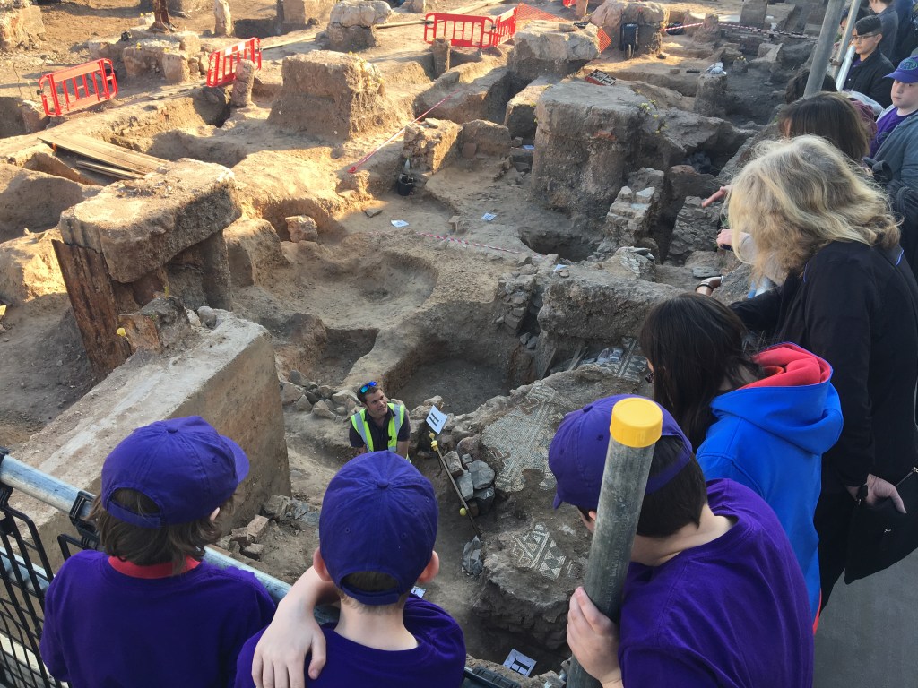 Children looking down from a viewing platform at a Roman mosaic as an archaeologists explains what they can see.